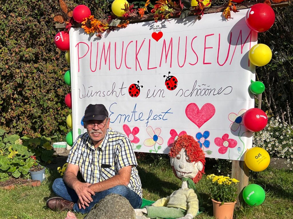 Pumuckl Museums Inhaber Sven Coorßen sitzt vor einem großen weißen Schild mit der Aufschrift PUMUCKLMUSEUM wünscht ein schööönes Erntefest. Das Schild ist ringsherum mit Luftballons geschmückt. Neben ihm eine Pumuckl Figur aus Stoff.