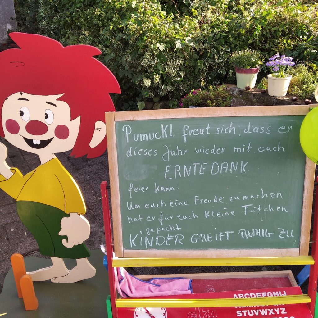 Pumuckl aus Holz Aufstellfigur neben einer Kinder Schultafel. Auf der Tafel steht "Pumuckl freut sich, dass er dieses Jahr wieder mit euch ERNTEDANK feiern kann. Um euch eine Freude zu machen hat er für euch kleine Tütchen gepackt. KINDER GREIFT RUHIG ZU.