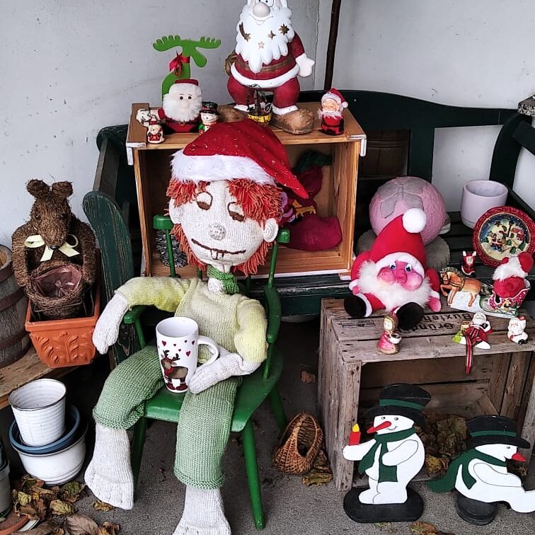 Pumuckl Stofffigur auf Stuhl sitzend mit Weihnachtsmütze auf und Porzellantasse in der Hand, umgeben von Weihnachtsdekorationen